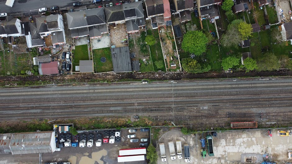 An aerial view of a residential garden area separated by multiple narrow garden plots, each with different planting arrangements and lawn textures. The gardens feature a variety of trees, shrubs, and