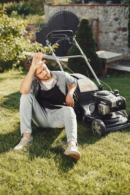 A man with light hair and a beard, dressed in casual grey pants, a checkered shirt, and a black apron, is sitting on a lush green lawn in a garden. He appears tired, with his left hand resting on his 